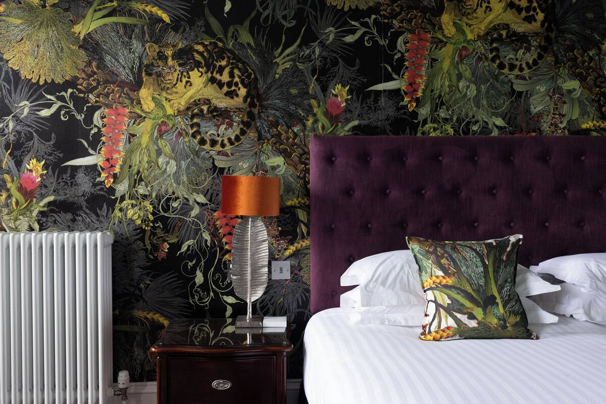 A bedroom with a large bed, a purple headboard, a nightstand with a lamp, and a floral wallpaper.