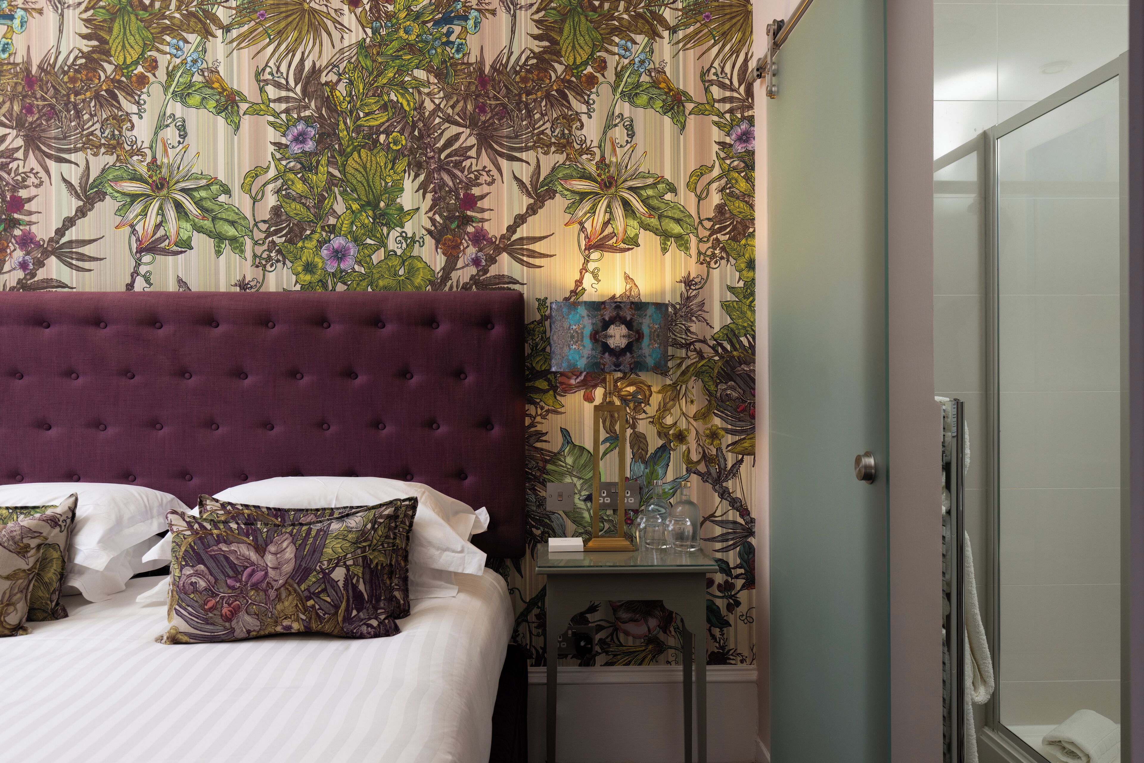 A bedroom with a tufted headboard, floral wallpaper, a bedside table with a lamp, and a glass shower enclosure.