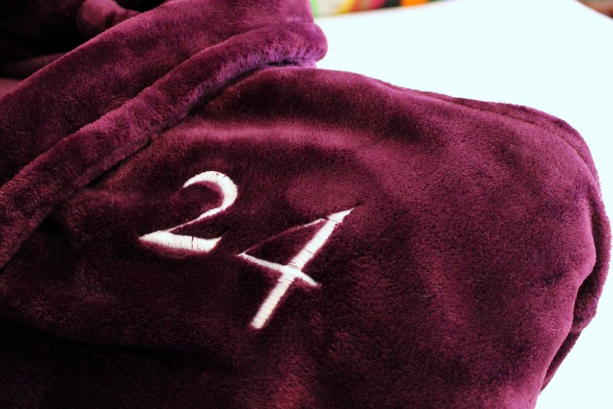 A plush, dark-colored robe with the number "24" embroidered on it, laid over a bed with a colorful, striped pillow and a tufted headboard.