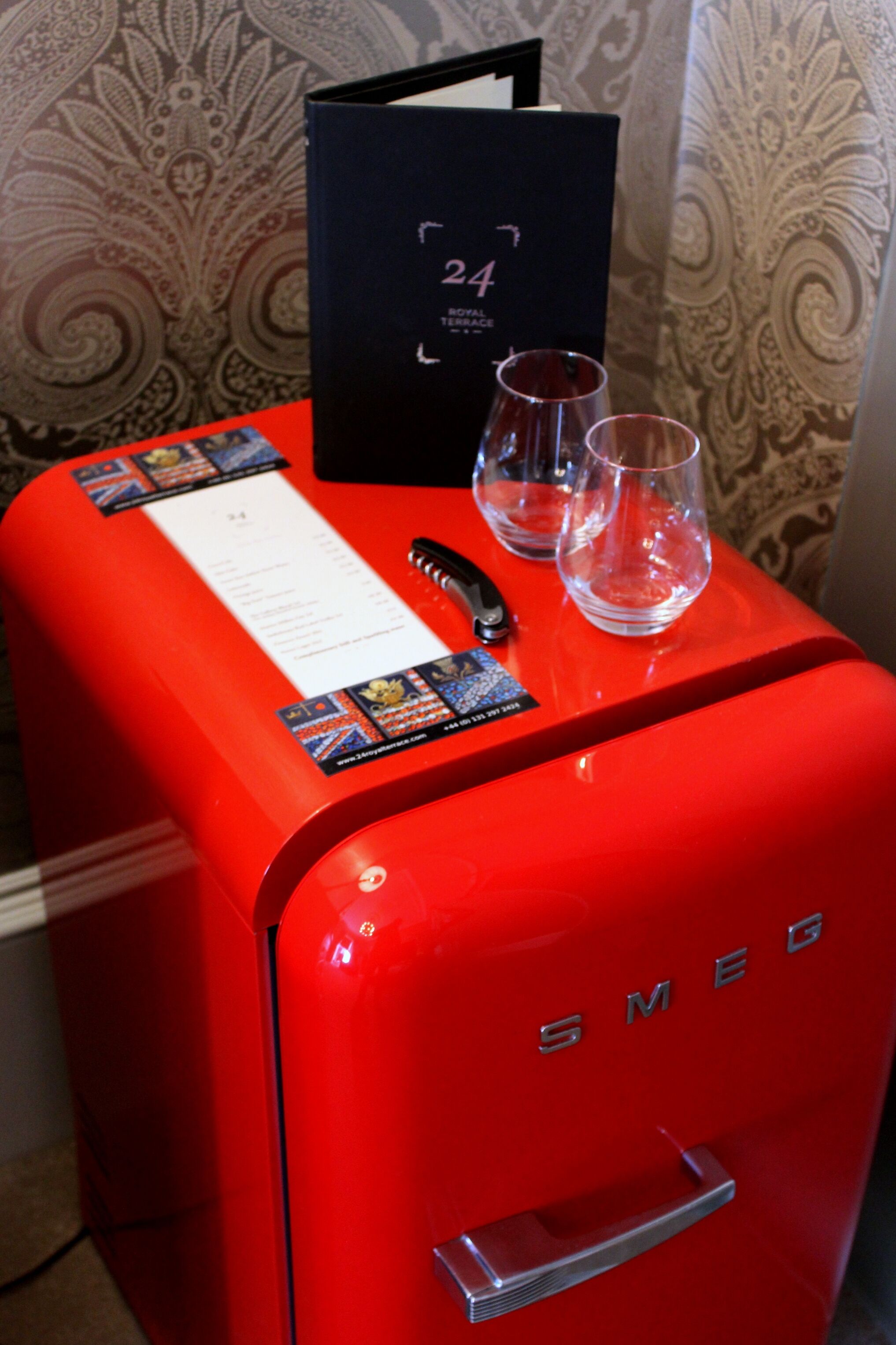 Una maleta roja con la etiqueta "SMEG", colocada sobre una superficie con una carpeta negra y dos vasos vacíos.