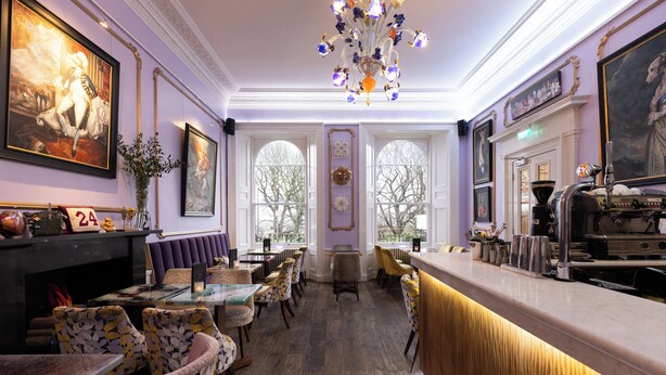 Un espace bar doté d’un long comptoir, de tabourets et d’une cheminée. Les murs sont ornés de tableaux encadrés et un lustre est suspendu au-dessus du bar.