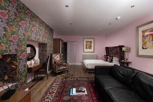 A room with a floral wallpaper, a black leather sofa, a bed, a desk with a chair, and a radiator.