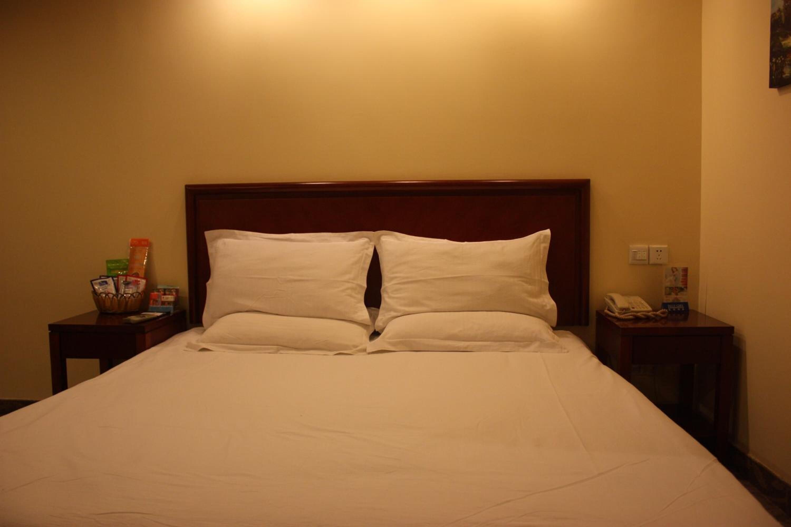 Une chambre d’hôtel avec un grand lit, deux oreillers, une tête de lit en bois, une table de chevet avec un téléphone et une lampe, et une petite table sur laquelle se trouve un panier rempli de divers objets.