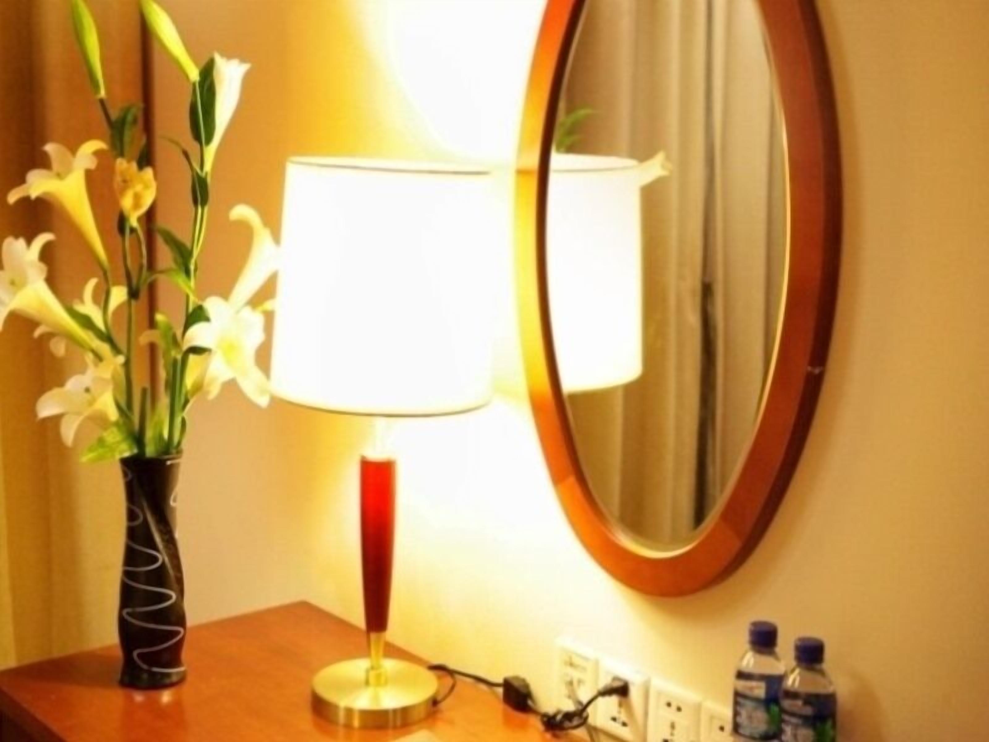 A lamp with a white shade and a red base, a vase with white flowers, and a mirror on a wooden surface.