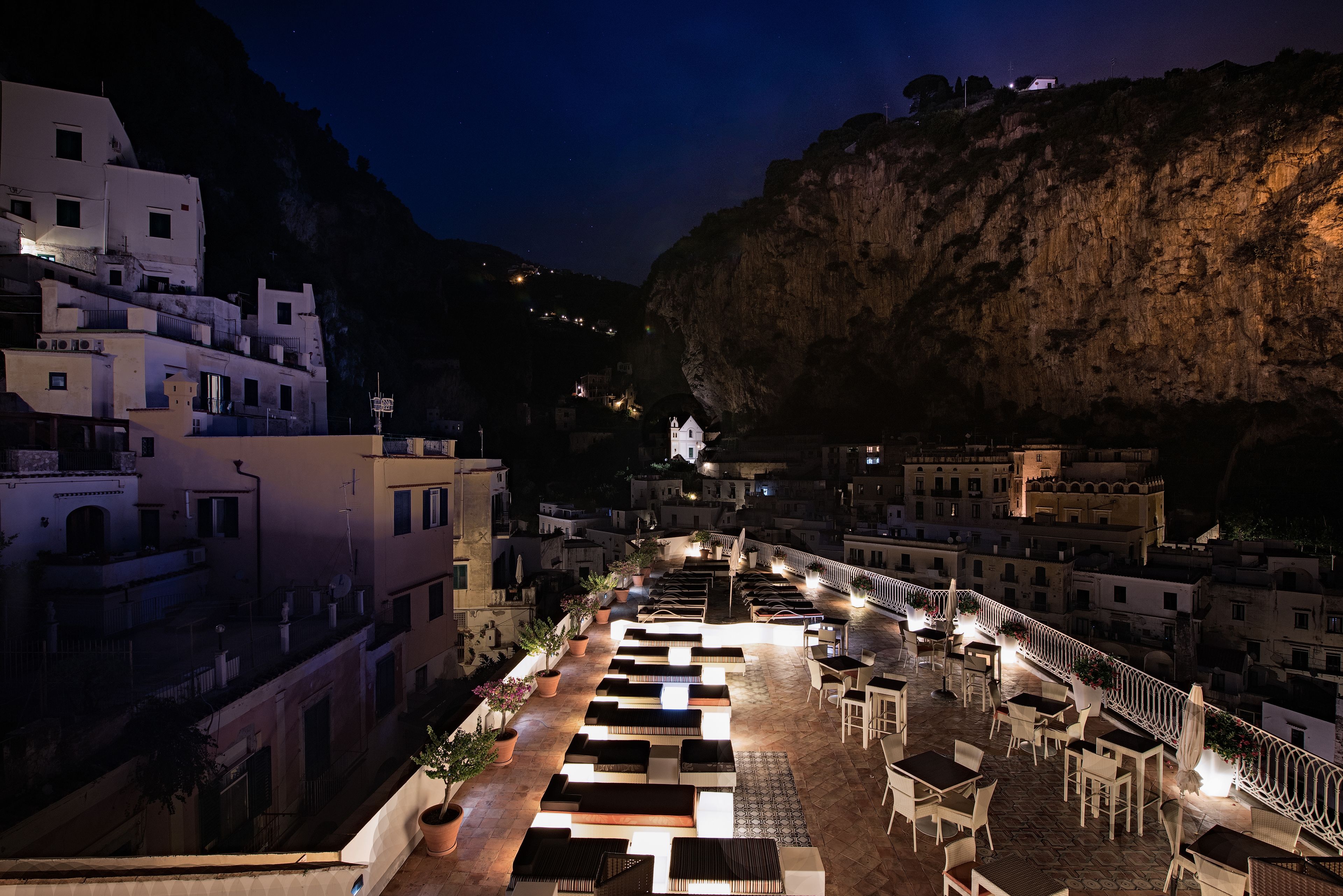A rooftop terrace with outdoor seating and a view of a coastal town at night.