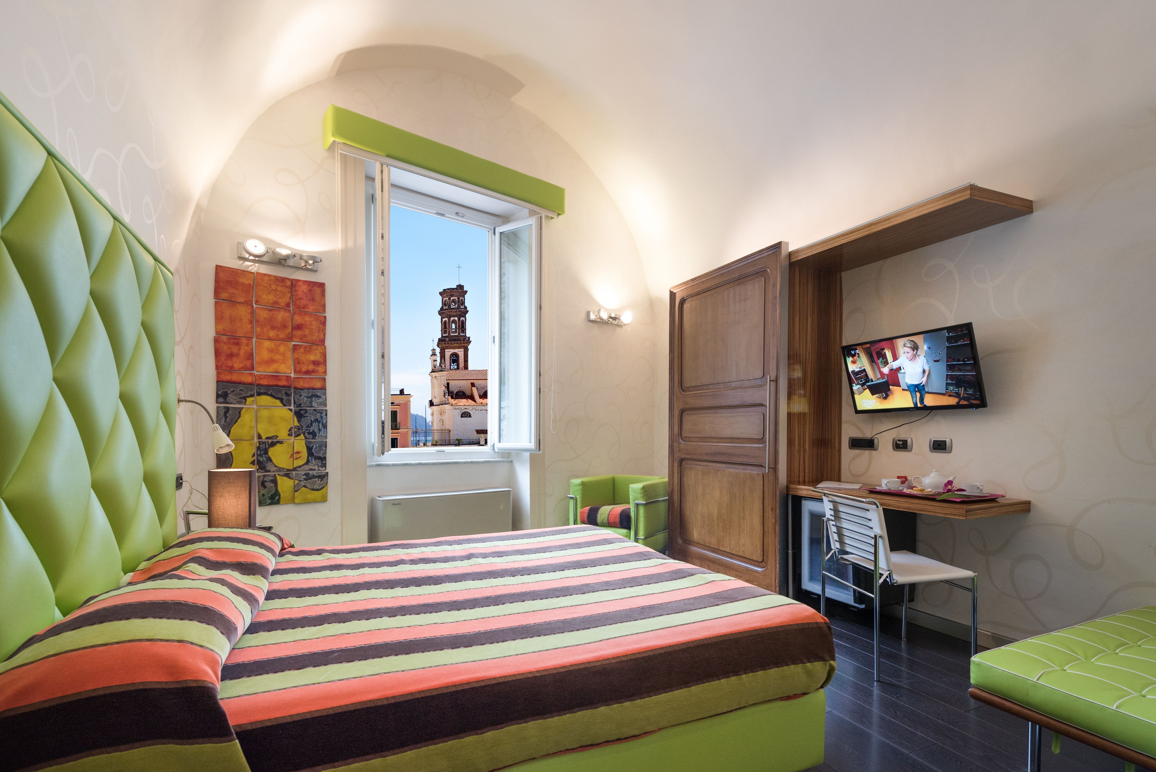 A hotel room with a bed, a desk, a television, a window with a view of a tower, and a green upholstered bench.