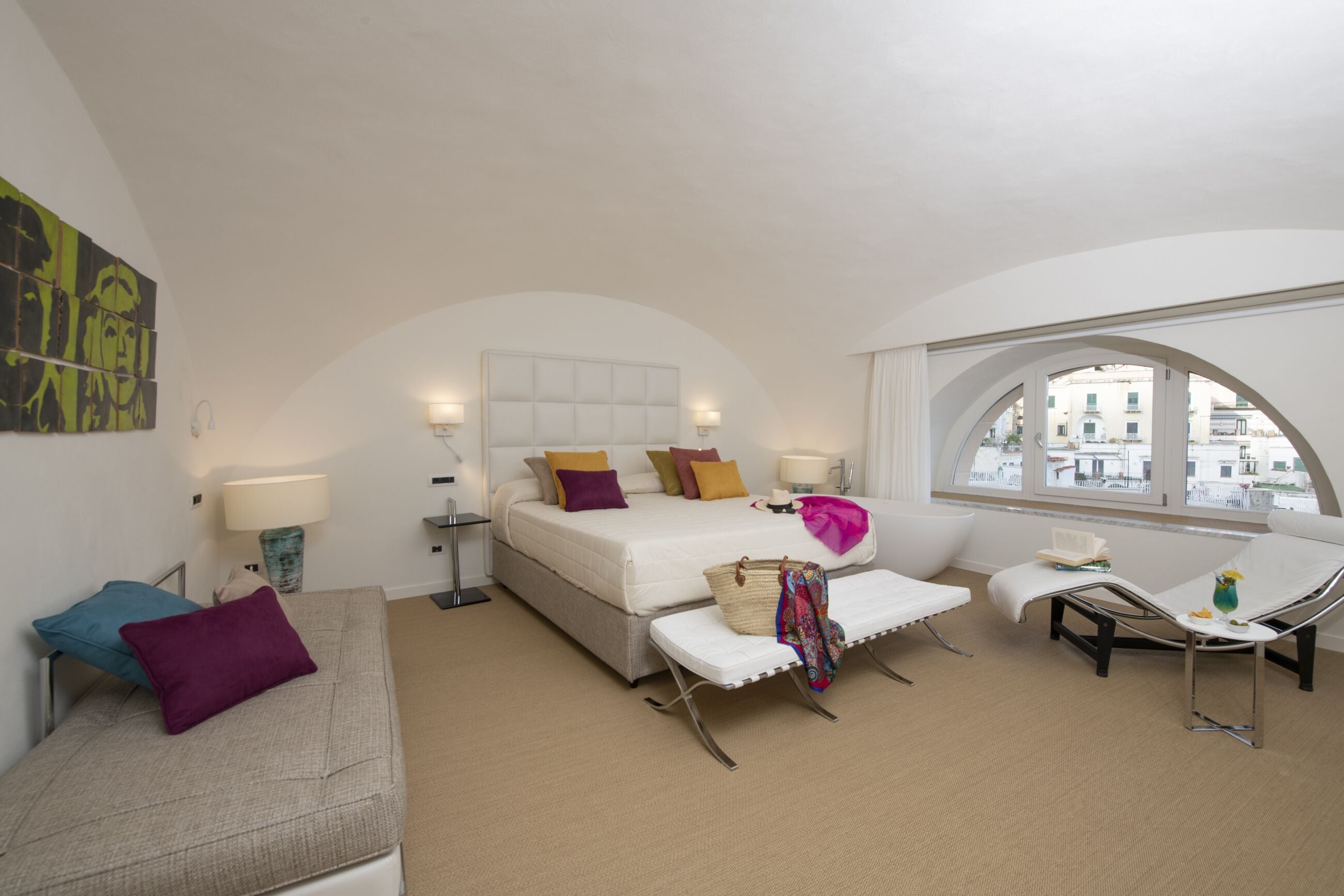 A modern hotel room with a large bed, a sofa, a chaise lounge, and a view of buildings through a large arched window.