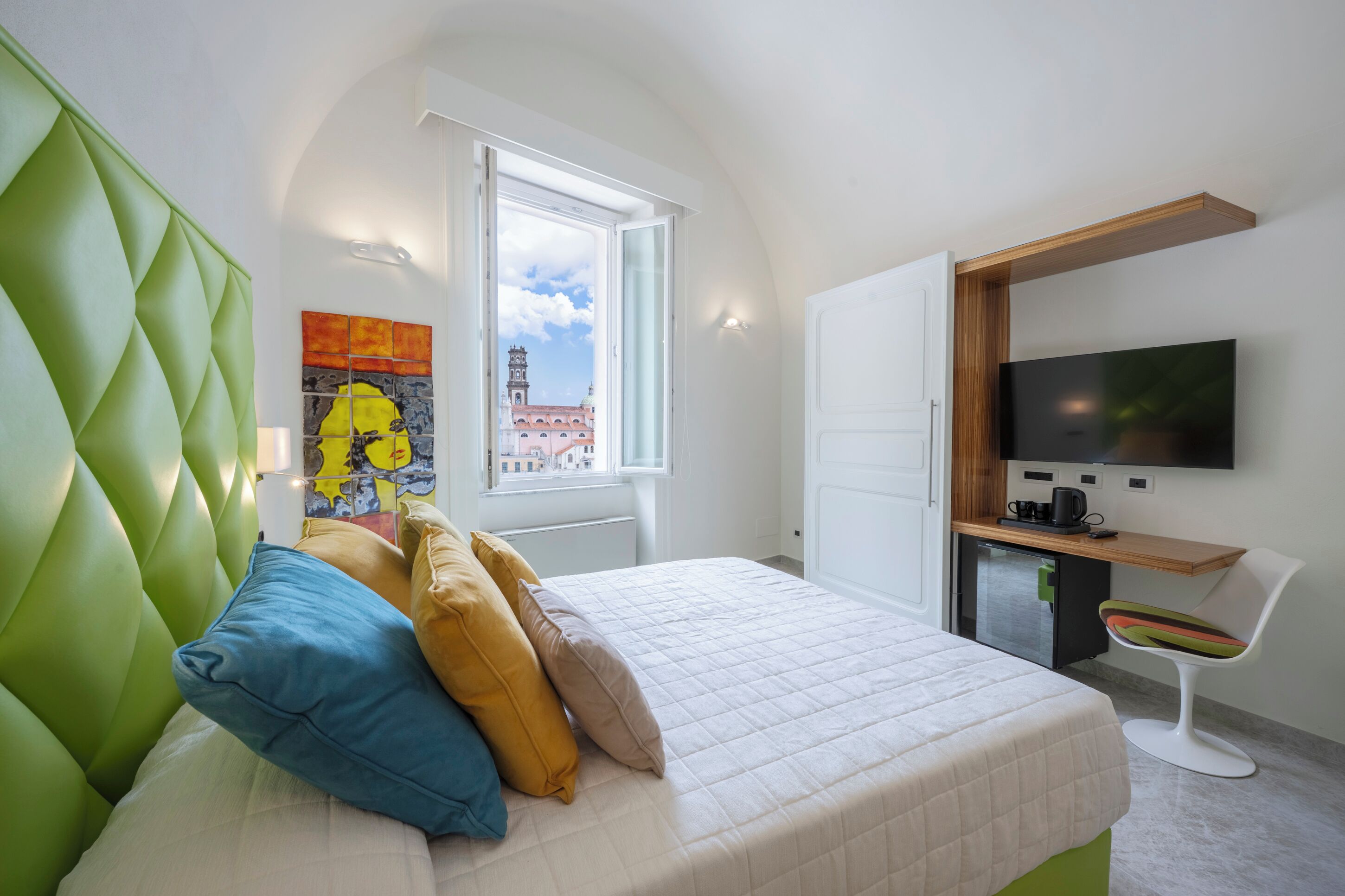 A modern hotel room with a green upholstered headboard, a bed with multiple pillows, a desk with a flat-screen TV, and a window with a city view.