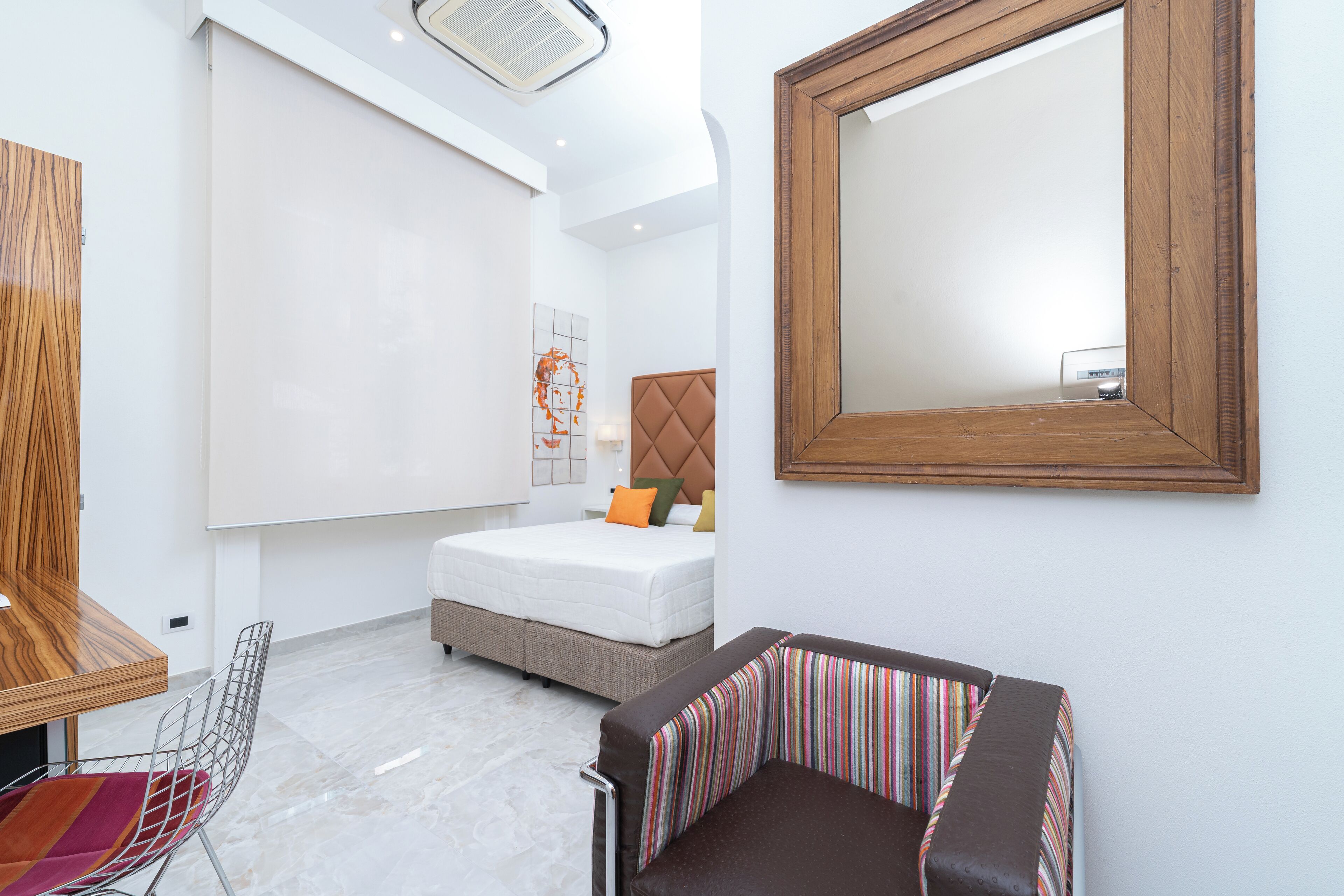 A modern hotel room with a bed, a sofa, a chair, a wooden table, and a large mirror.