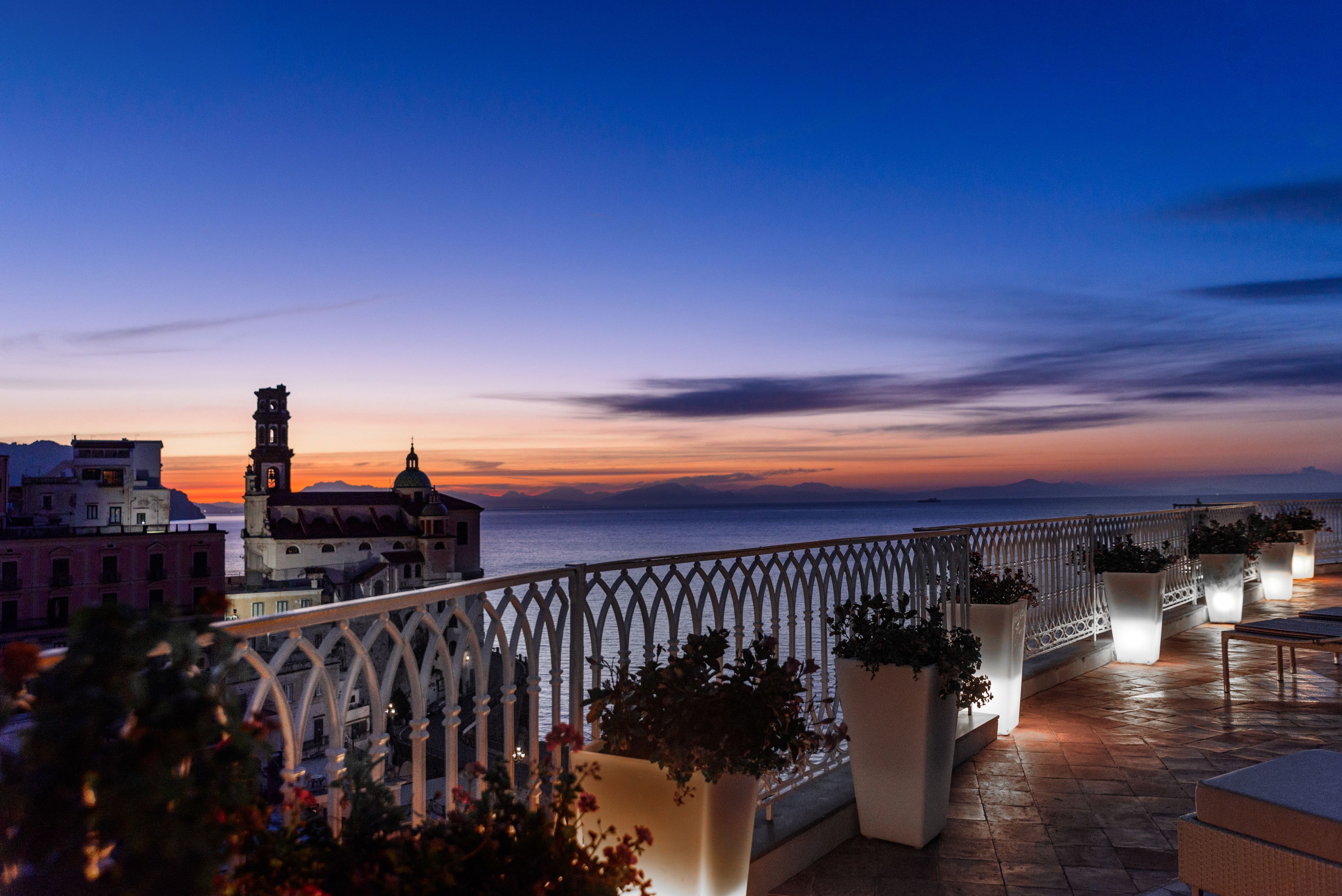 Un balcon agrémenté de plantes en pot, une balustrade et une vue sur la mer au coucher du soleil.