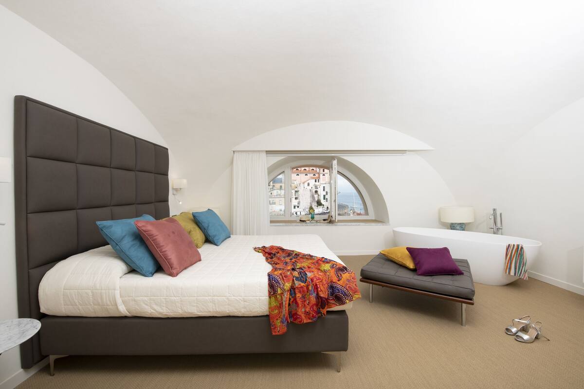Un dormitorio con una cama grande, una banca, una bañera y vistas a edificios a través de una ventana.
