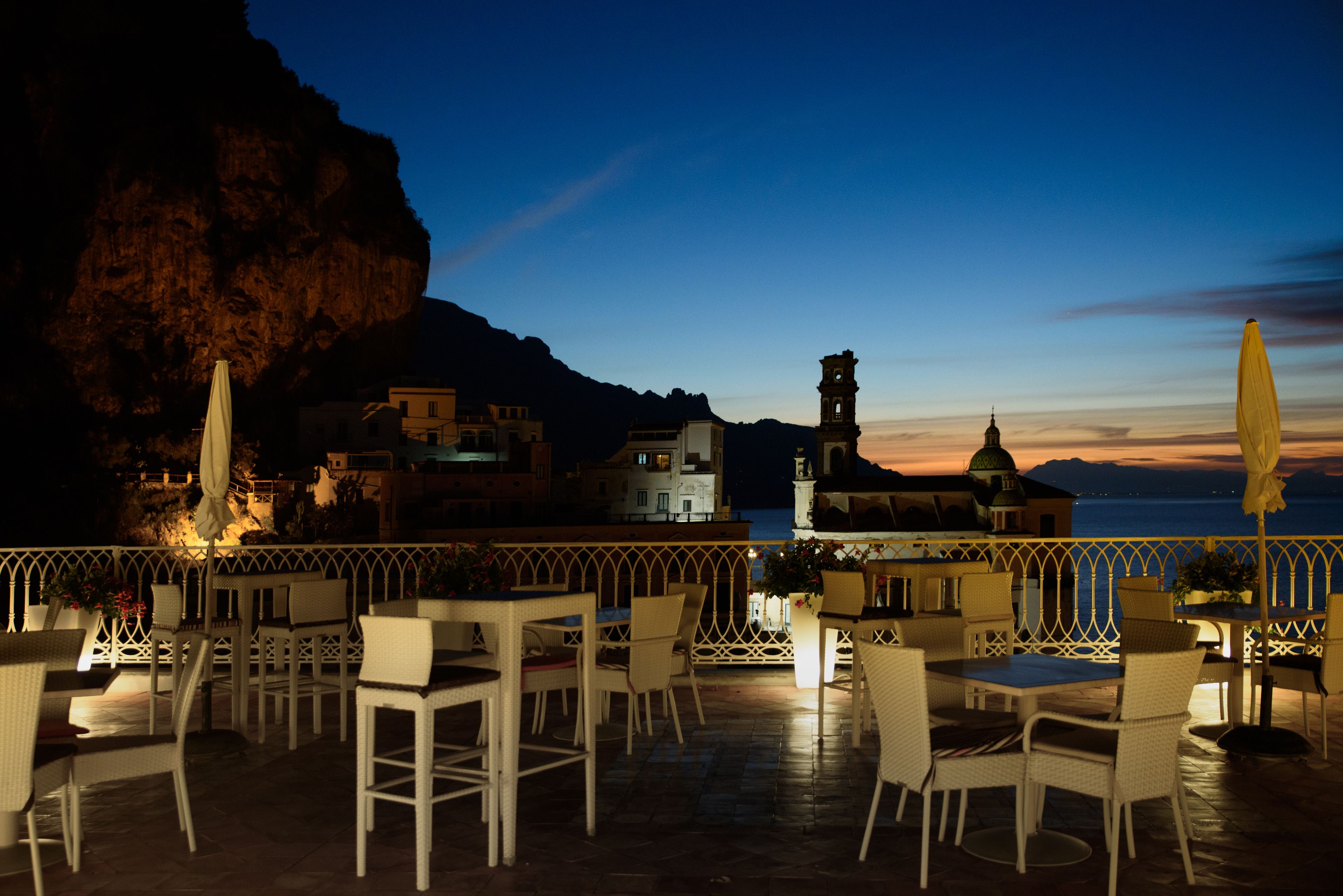 A terrace with outdoor seating and a view of a coastal town at dusk.