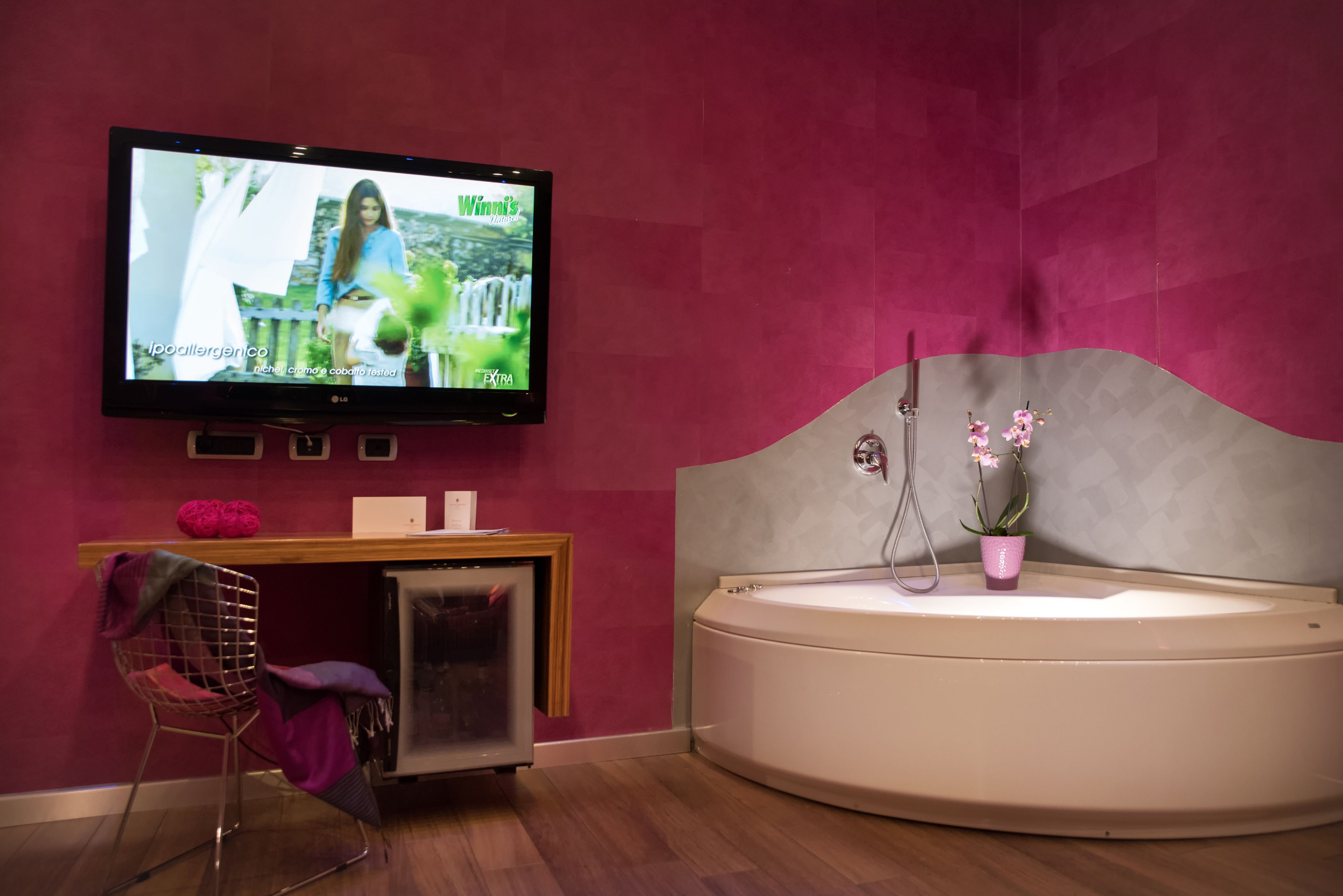 Ein modernes Badezimmer mit einem großen Fernseher an der Wand, der eine Sendung zeigt, einer Badewanne mit einer rosa Pflanze und einem hölzernen Waschtisch mit einem rosa Stuhl.