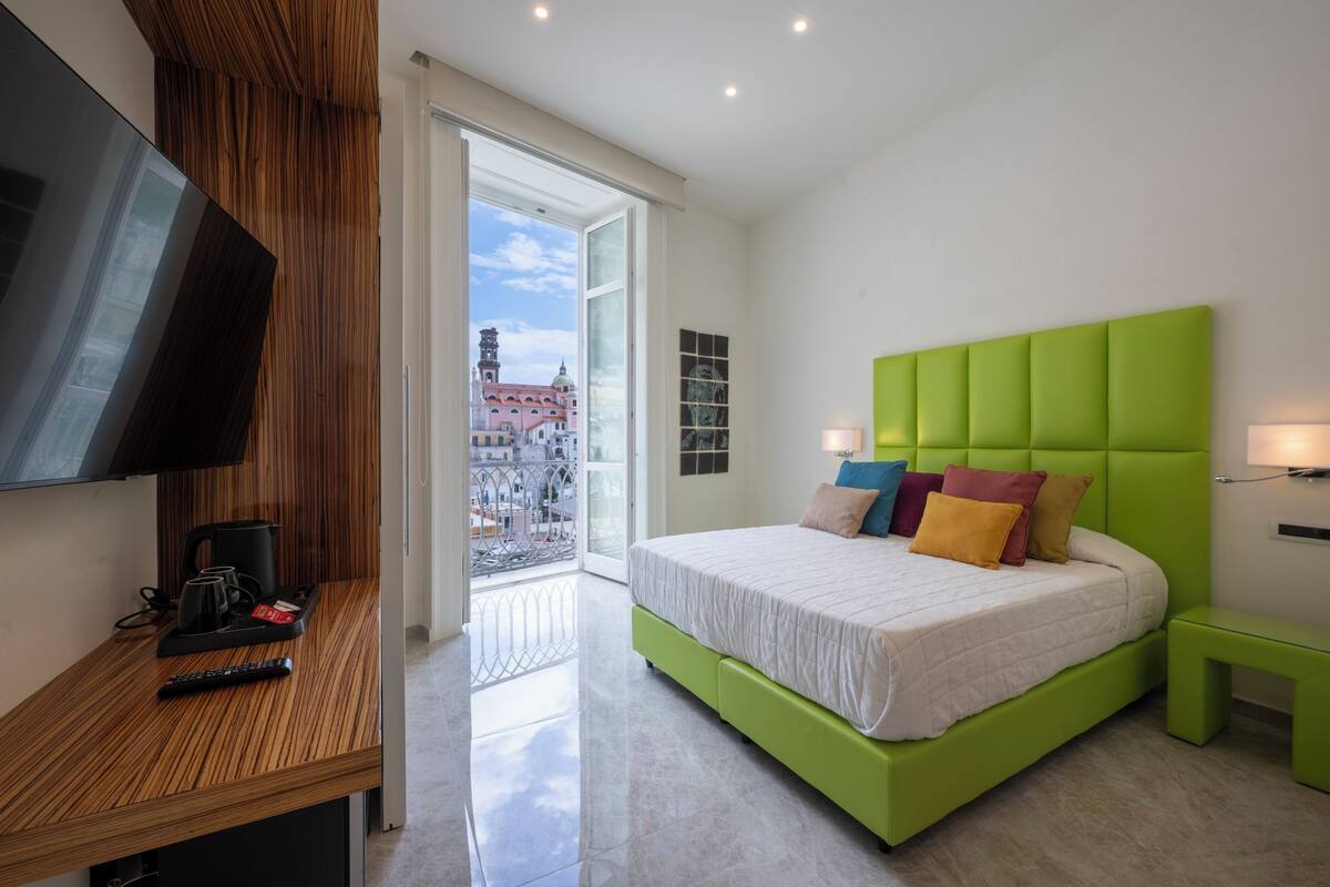 Habitación de hotel moderna con una cama verde, un televisor, un balcón con vista a la ciudad y un escritorio de madera con cafetera.