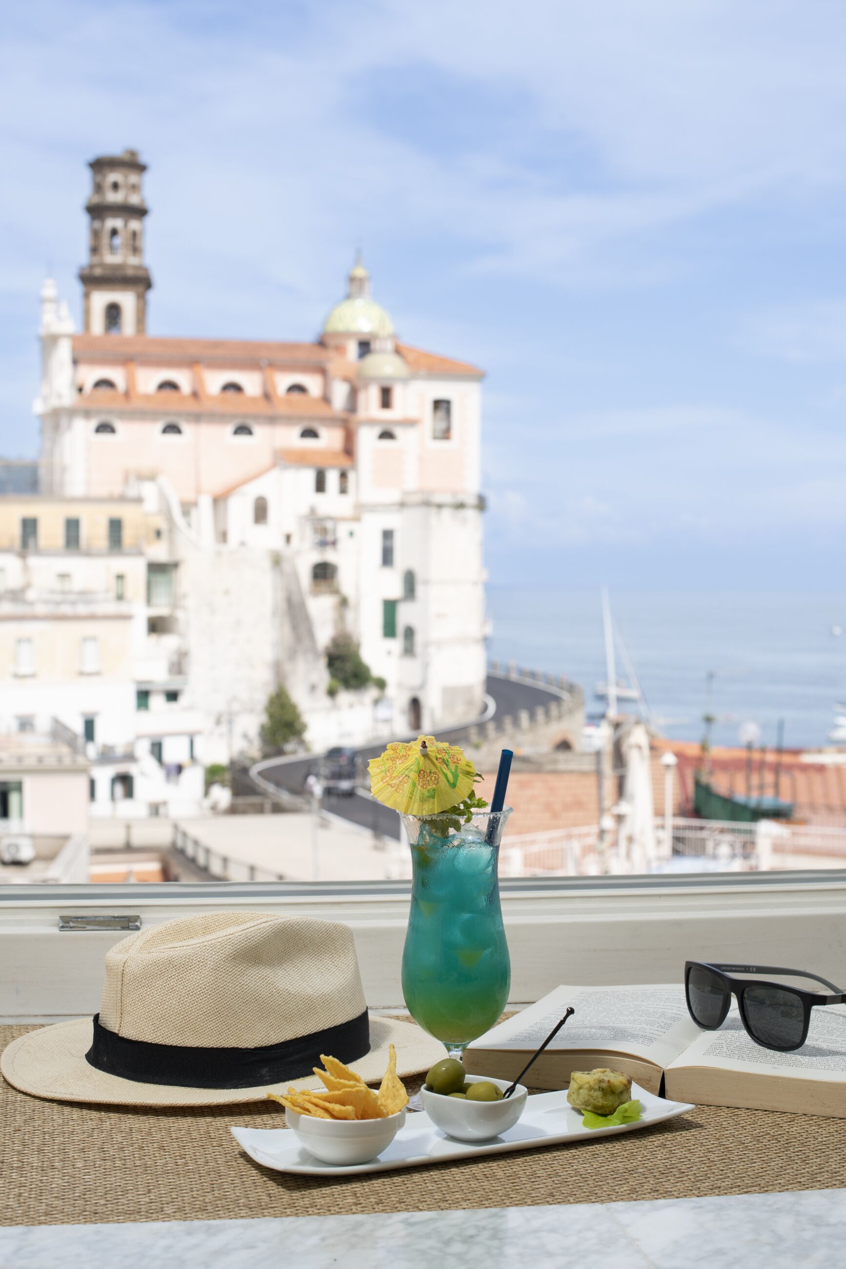 Una mesa con un sombrero, una bebida, aperitivos y gafas de sol, con vista a un edificio histórico y al mar de fondo.