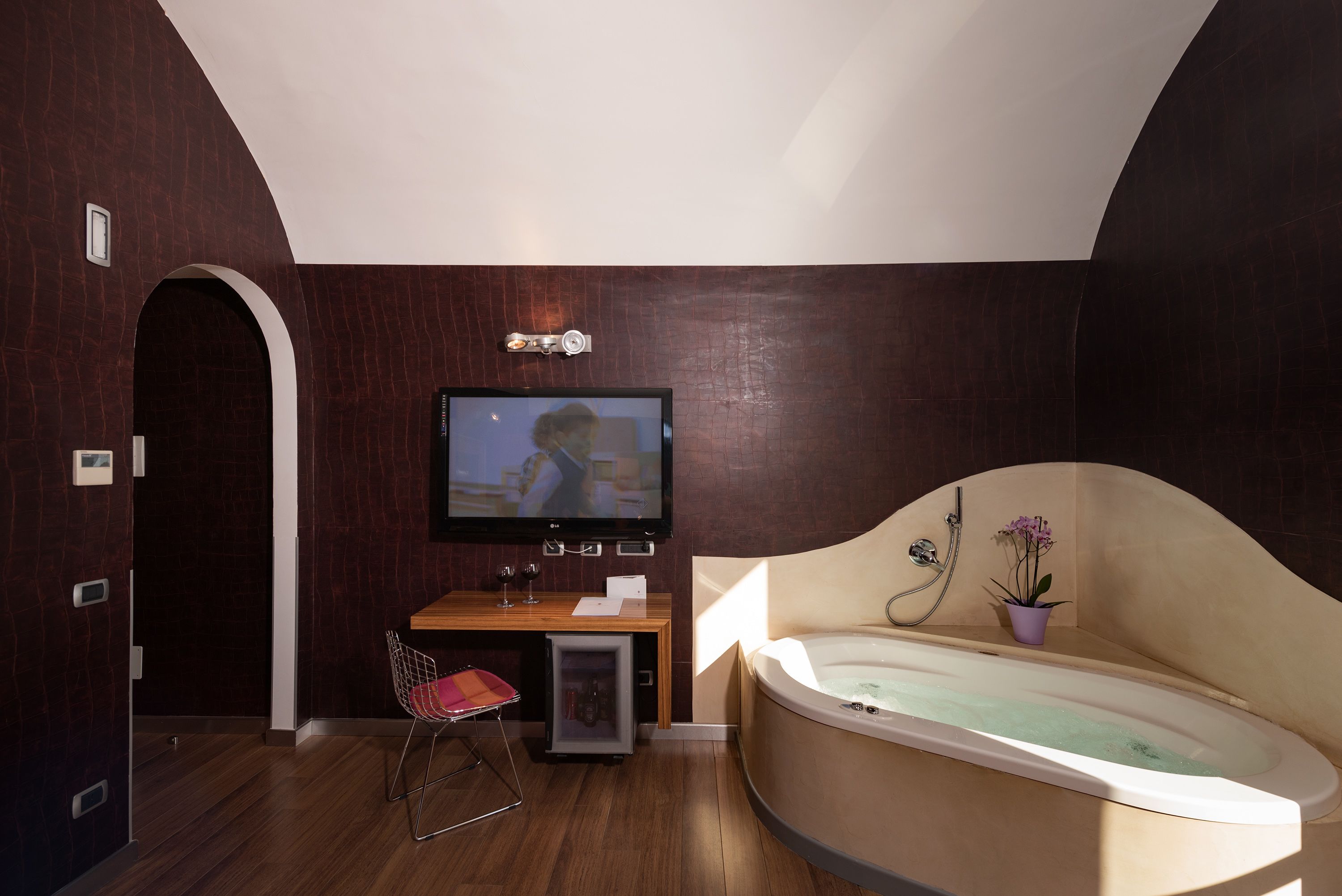 A modern bathroom with a large bathtub, a television mounted on the wall, and a small seating area with a chair and a table.