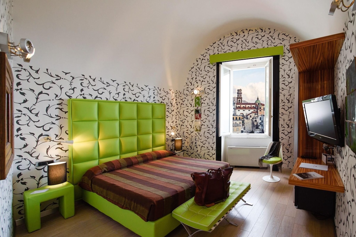 Una habitación con una cama verde, televisión, una mesita pequeña y un banco.