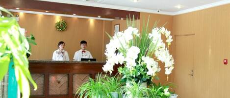Ein Hotel-Empfangsbereich mit einem Holzbüro, zwei Mitarbeitern und einem Glastisch mit weißen Blumen und Topfpflanzen.