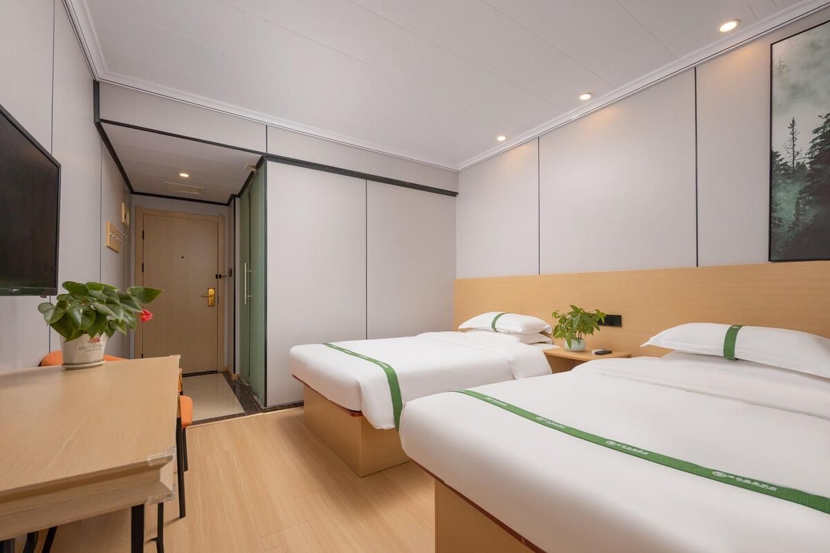 A hotel room with two beds, a desk, and a chair. There is a plant on the desk and another on the bedside table.