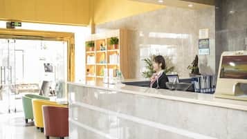 A hotel reception area with a receptionist, a reception desk, a seating area with colorful chairs, and a plant.