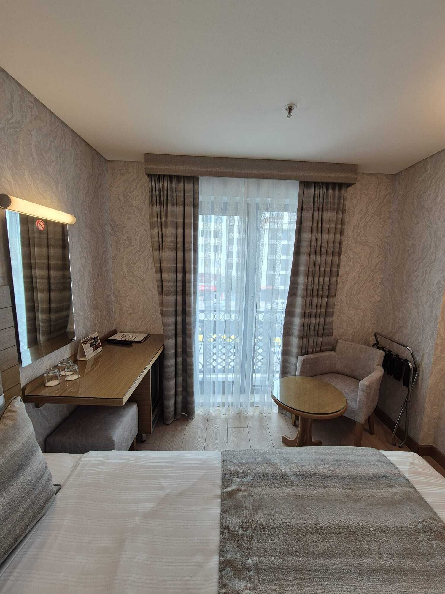 Habitación de hotel con una cama grande, un sofá, una mesita y un escritorio. Se ve una ventana con cortinas y vistas a edificios.
