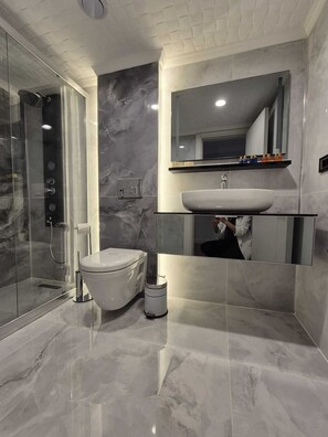 Une salle de bain moderne avec une douche à l’italienne en verre, des toilettes blanches, un sol carrelé en marbre et un grand miroir au-dessus d’un lavabo.