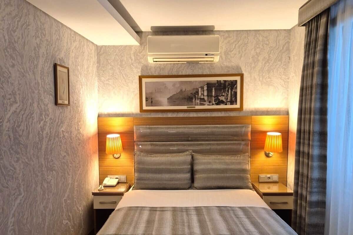 A hotel room with a large bed, two bedside tables with lamps, a mounted air conditioner, and a framed picture on the wall.