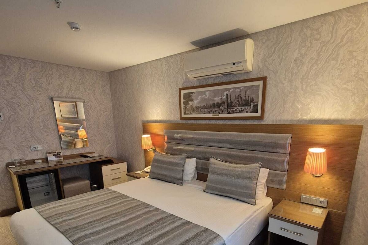 A modern hotel room with a large bed, bedside tables with lamps, a desk, and a framed picture on the wall.