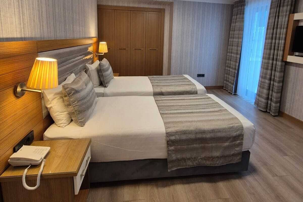 A hotel room with two beds, a wooden nightstand, a desk, a lamp, and a wardrobe.