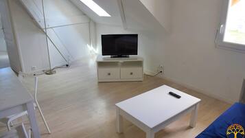 Habitació amb televisió sobre un moble blanc, una taula blanca i un llit blau.