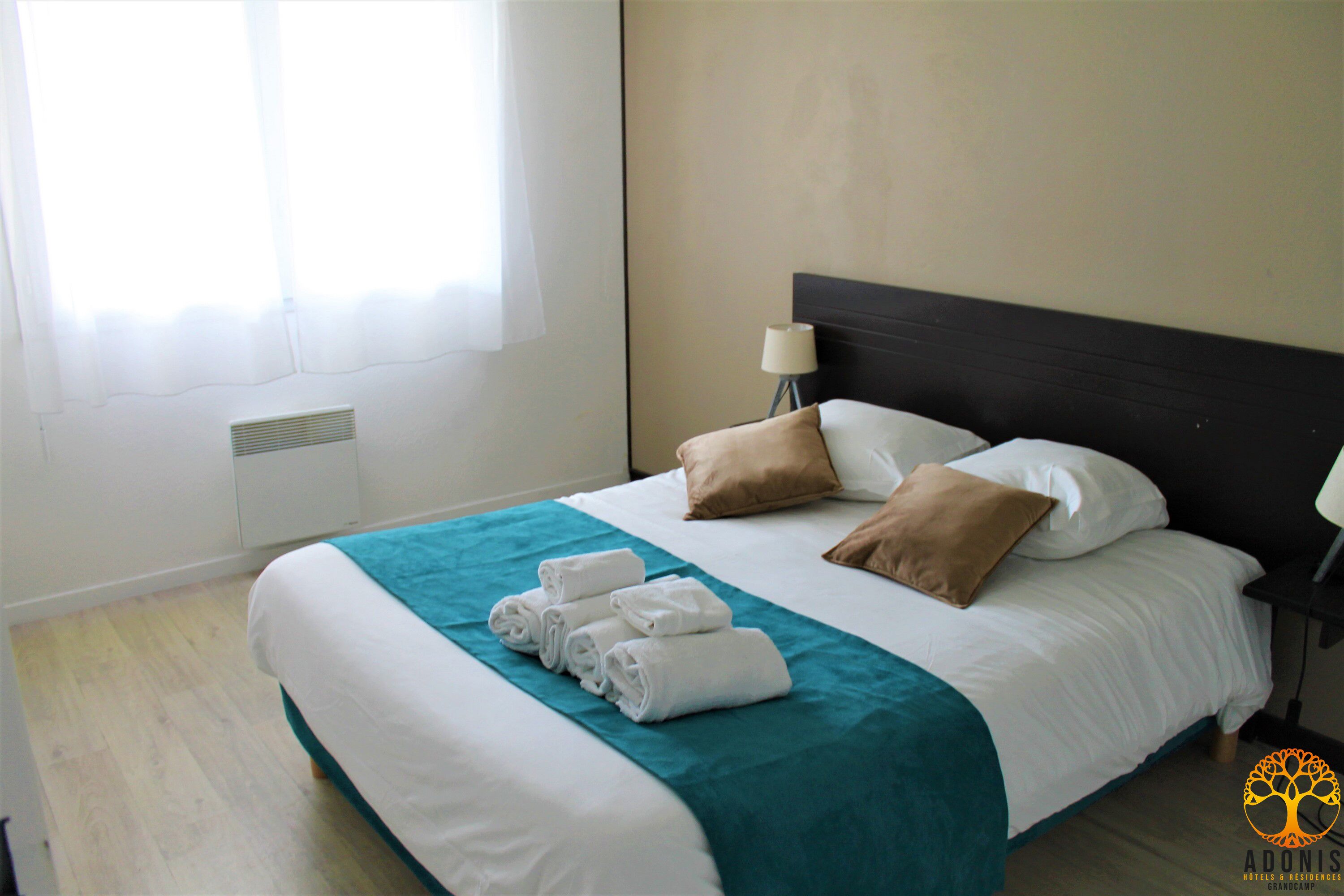 A neatly made bed with a teal bedspread, white linens, and two brown pillows. There are folded towels at the foot of the bed. A bedside lamp is on a nightstand to the right.