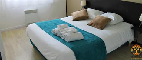 Una cama bien hecha con un edredón verde azulado, sábanas blancas y dos almohadas marrones. Hay toallas dobladas al pie de la cama. Una lámpara de noche está sobre una mesita a la derecha.