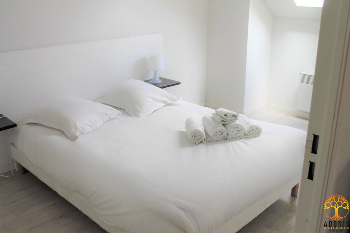 Un llit ben arrenjat amb llençols i tovalloles blancs, una taula de nit amb una làmpada, i un disseny d'habitació modern i minimalista.