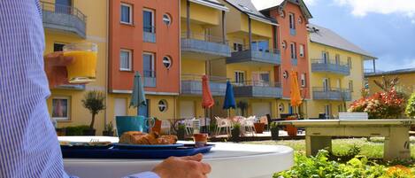 Una persona sosteniendo un vaso de jugo de naranja en una mesa con alimentos para el desayuno. El entorno es al aire libre, con un edificio amarillo al fondo que cuenta con balcones y mesas exteriores.
