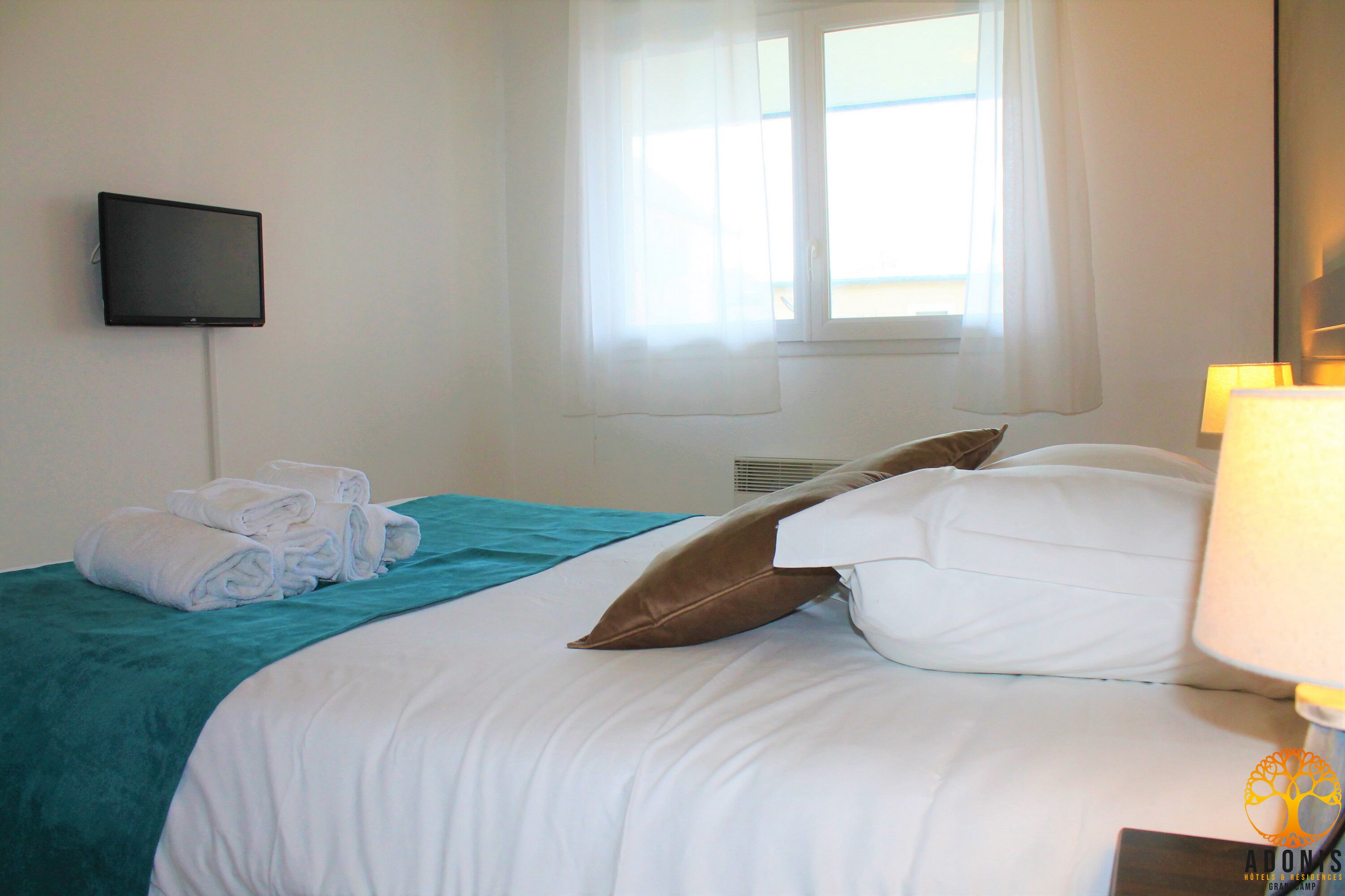 A hotel room with a bed, a flat-screen TV mounted on the wall, a window with curtains, and a bedside lamp.