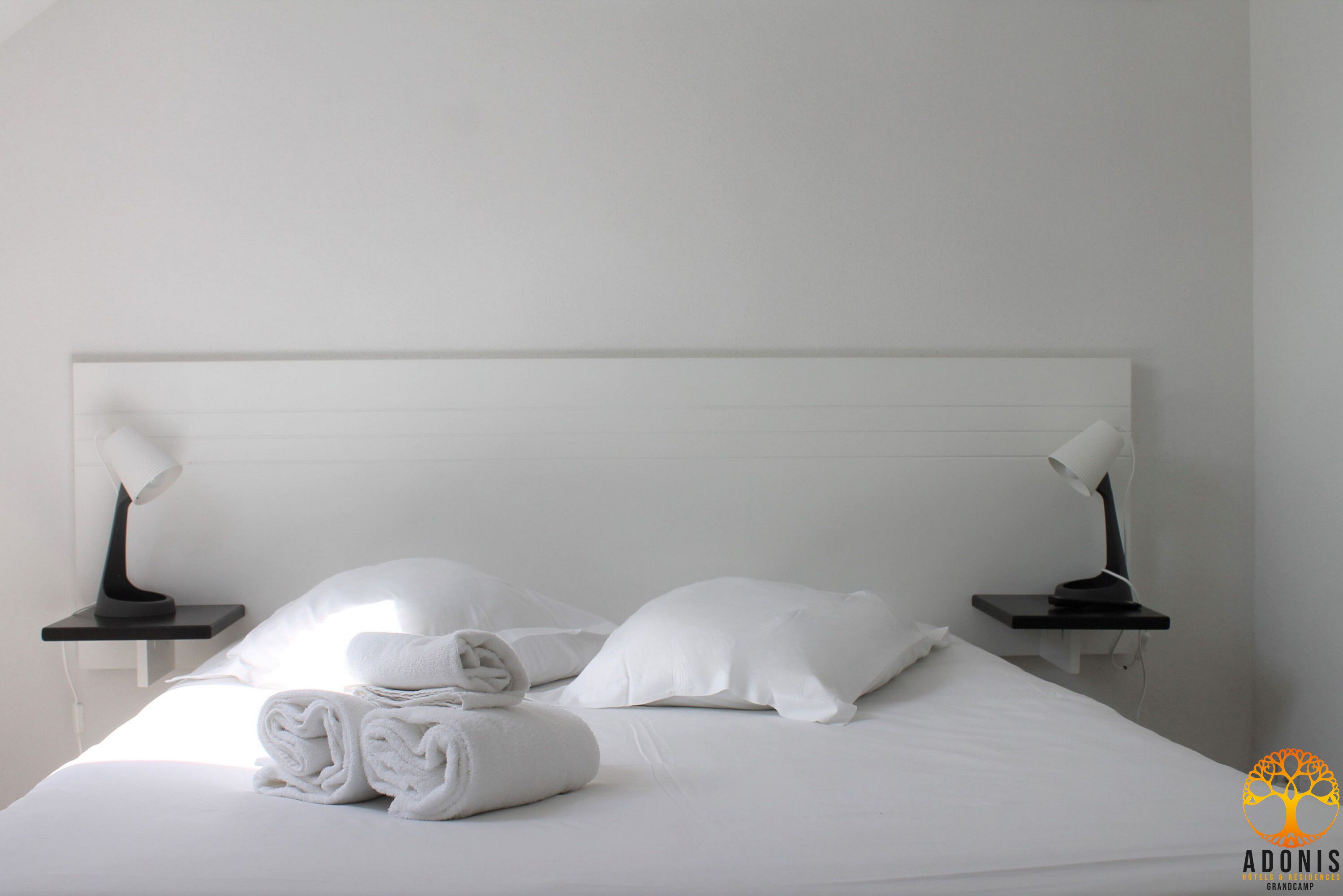 Un lit bien fait, avec du linge de lit blanc et des oreillers, deux tables de chevet avec des lampes, et une chambre au design épuré et minimaliste.