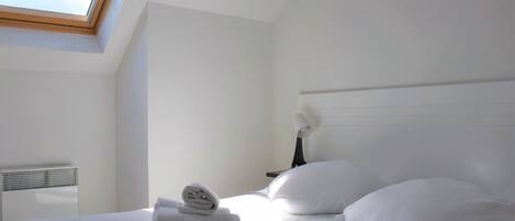A hotel room with a skylight, a bed with white linens, a bedside table with a lamp, and a wall-mounted heater.