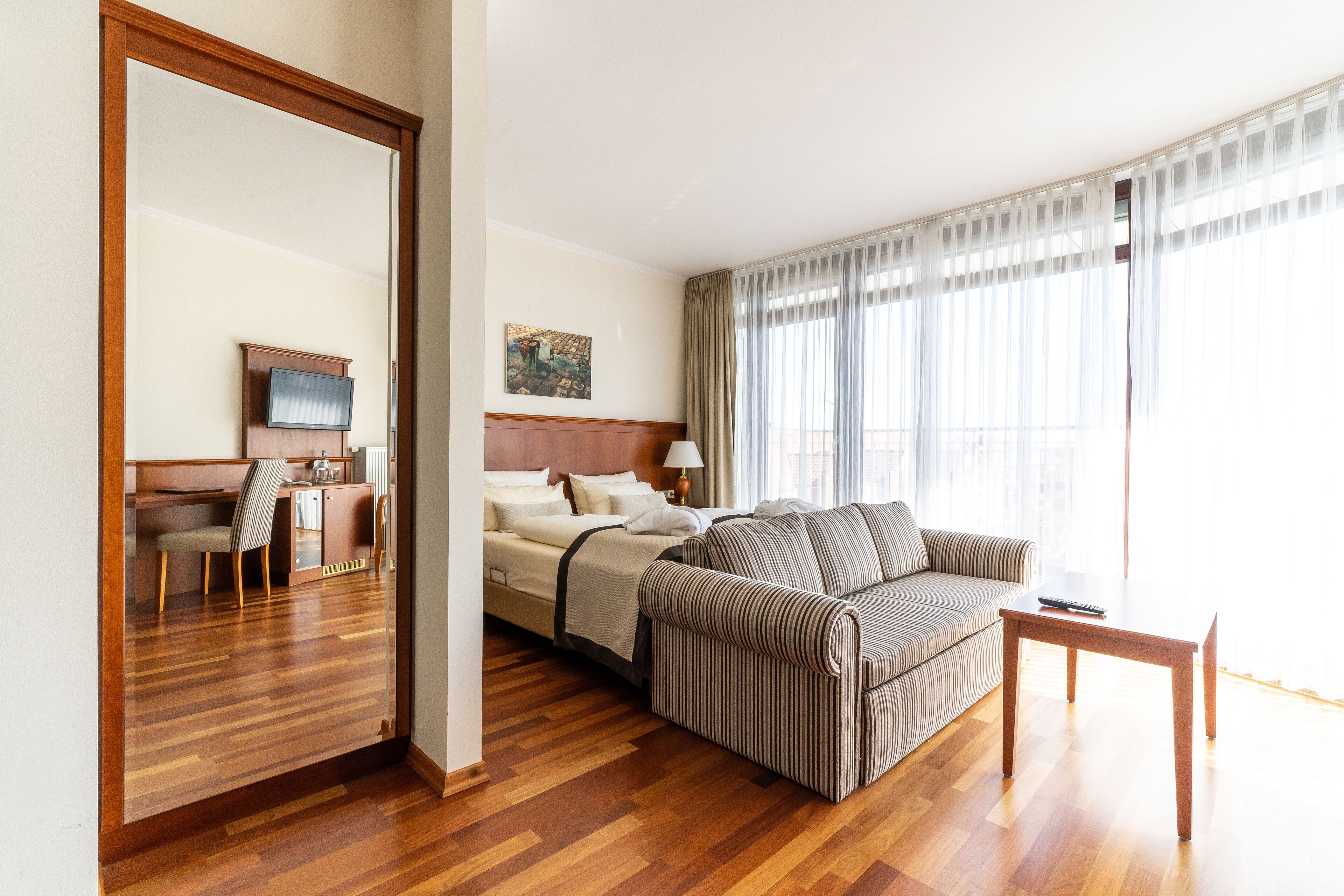 Ein Hotelzimmer mit Holzboden, einem Bett mit Kopfteil, einer gestreiften Couch, einem kleinen Tisch, einem Fernseher und einem Bild an der Wand.