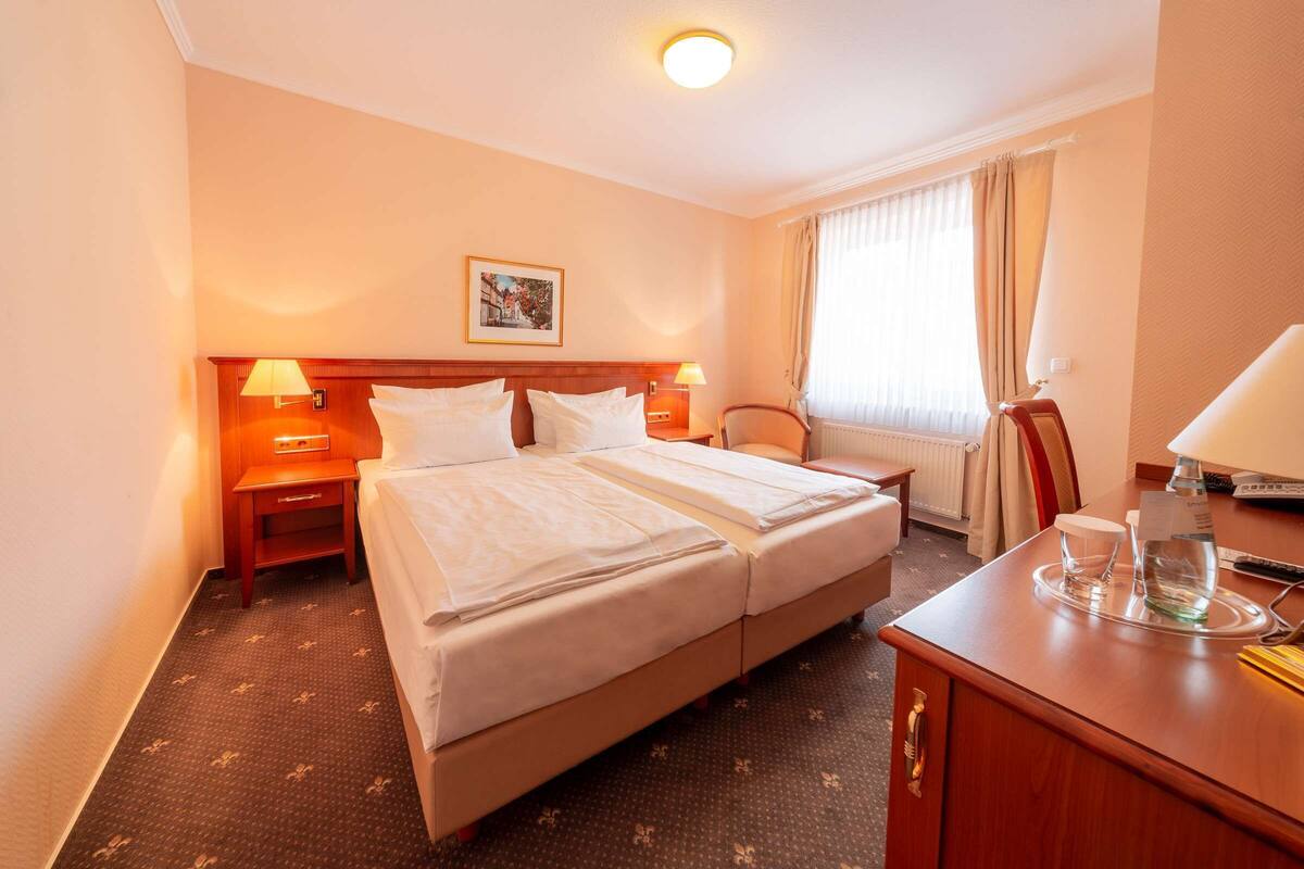 Een hotelkamer met een tweepersoonsbed, twee nachtkastjes met lampen, een houten bureau met stoel, een raam met gordijnen en een lijst met foto aan de muur.