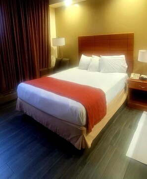 Habitación de hotel con una cama grande, cabecera de madera, mesita de noche con lámpara y cortina.
