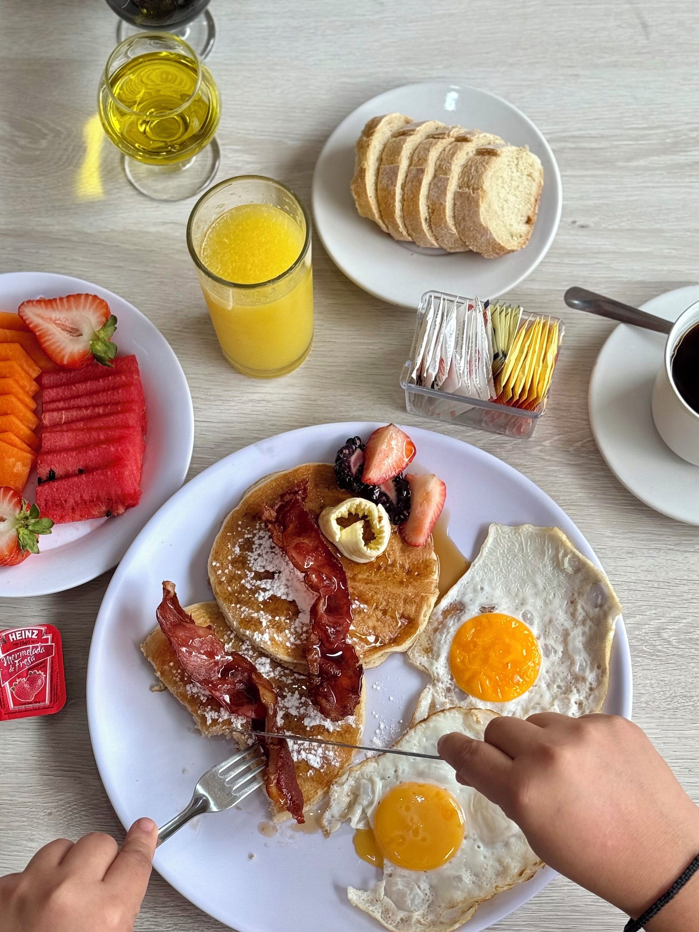 Un plato de desayuno con tortitas, tocino, huevos y fruta, un vaso de zumo de naranja y una taza de café sobre una mesa de madera.