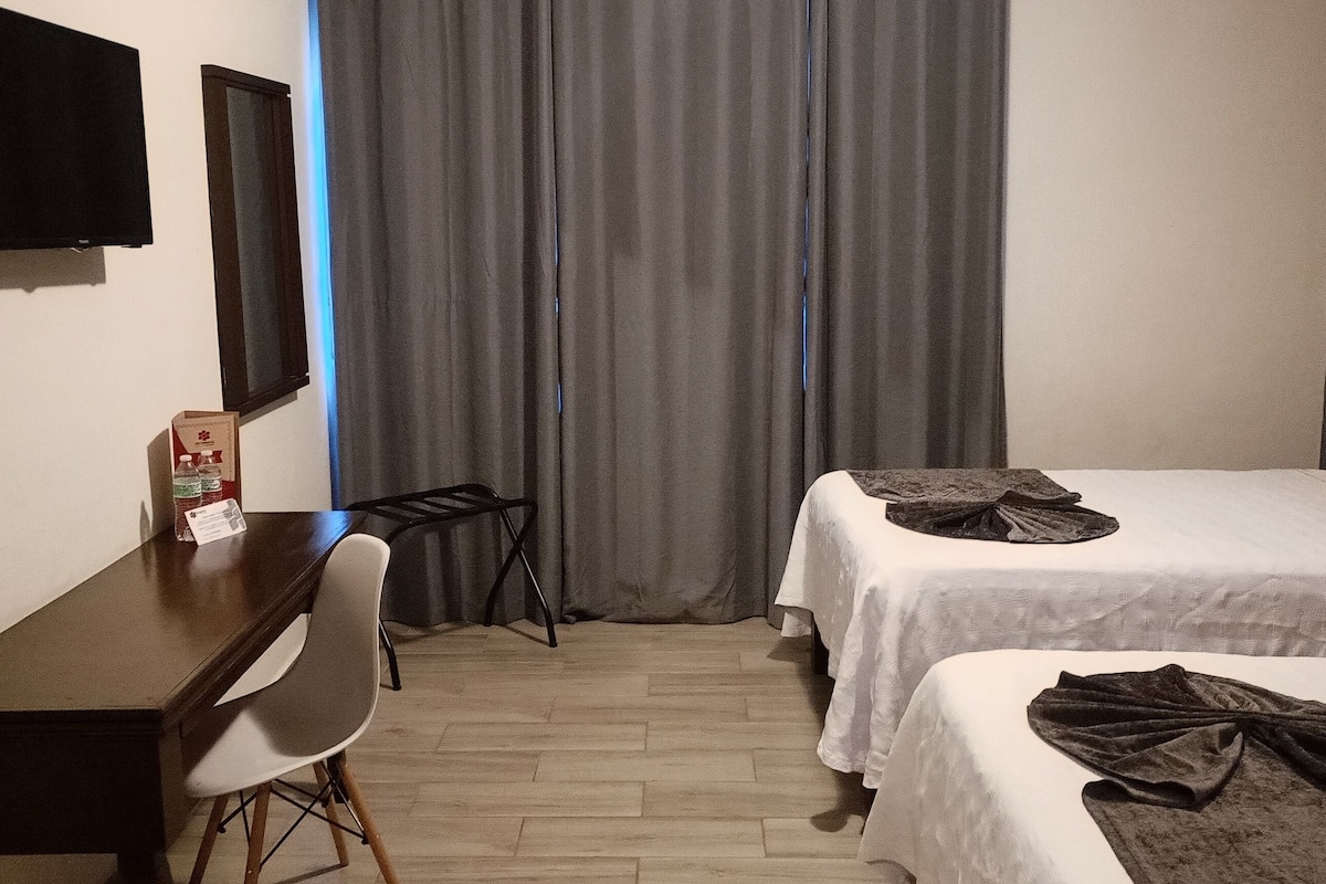 Habitación de hotel con dos camas, un escritorio, una silla, un televisor de pantalla plana y una ventana con cortinas.