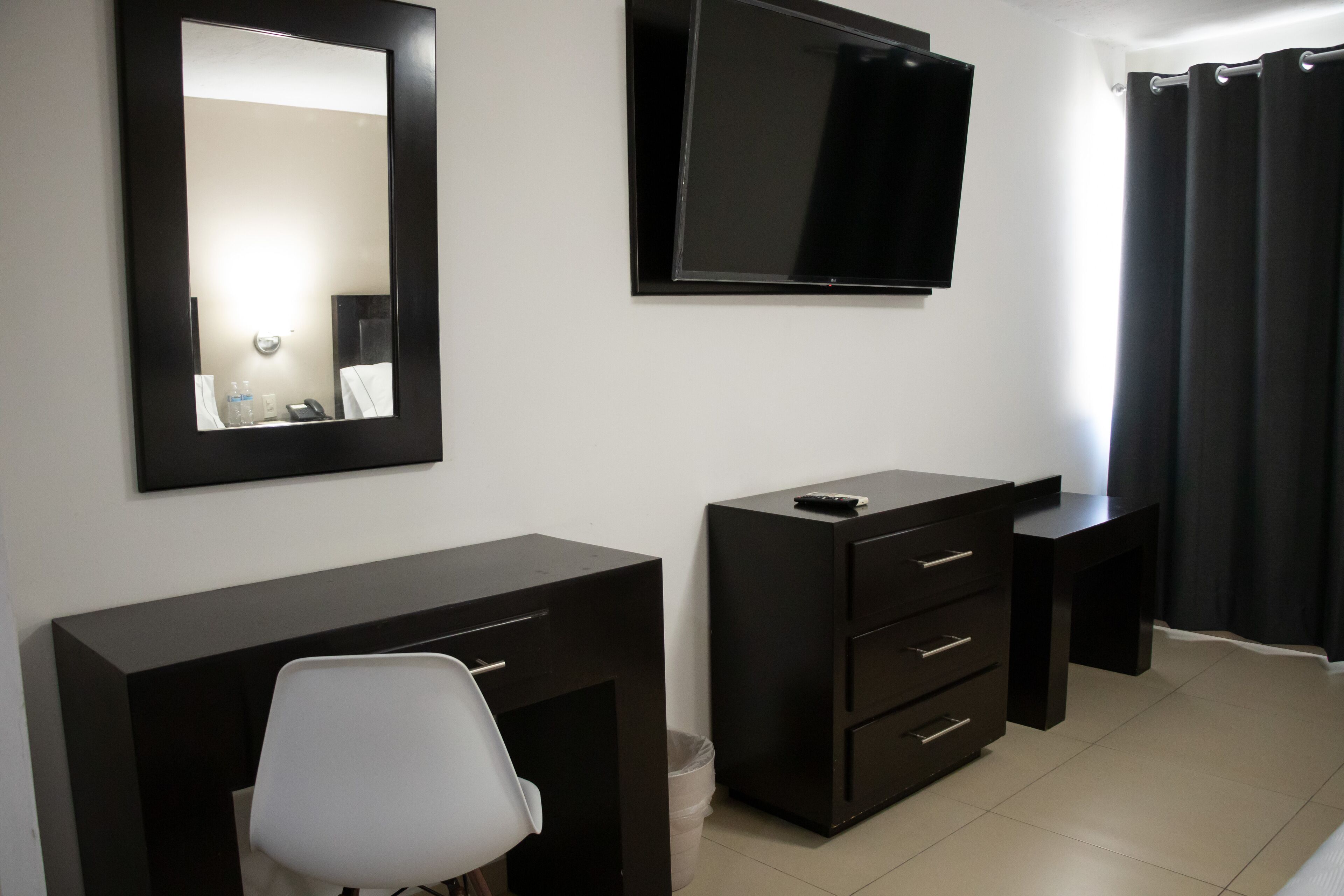 A hotel room with a large mirror, a flat-screen TV mounted on the wall, a dark-colored desk with drawers, a white chair, and a small trash bin.