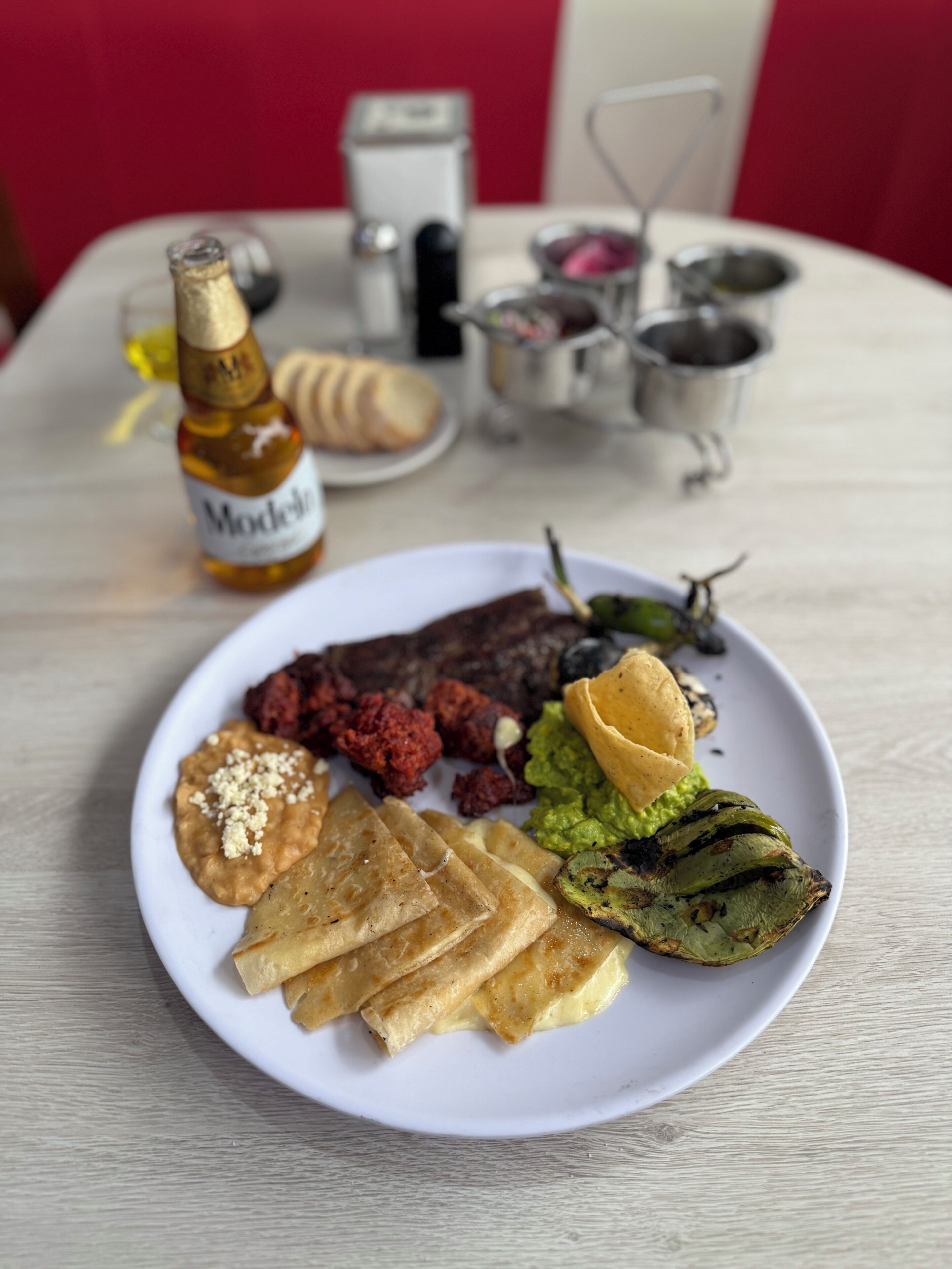A plate of food with a bottle of Modelo beer, a small plate with bread, and a metal container with condiments on a table.