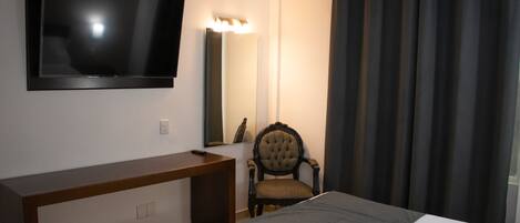 Una habitación de hotel con una televisión de pantalla plana montada en la pared, un escritorio de madera, un sillón tapizado y una cama con colcha gris.