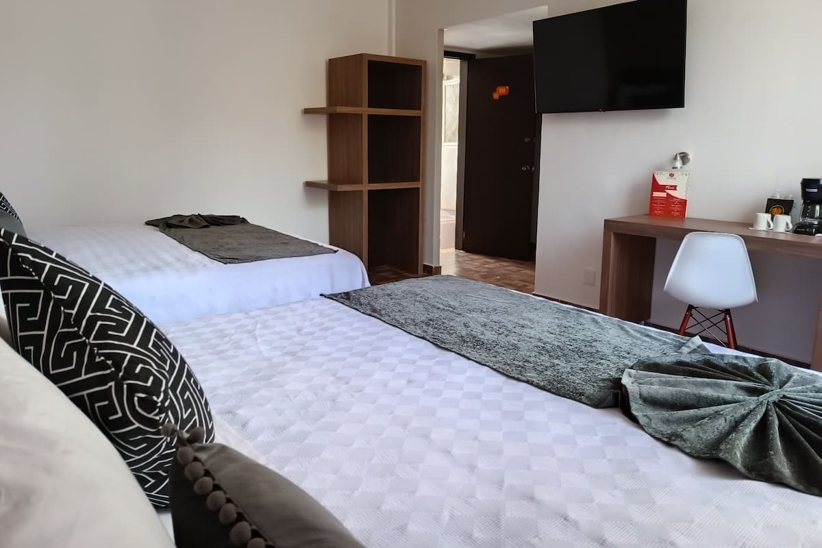 Habitación de hotel con dos camas, un escritorio con silla, televisor de pantalla plana y una pequeña zona de kitchenette.