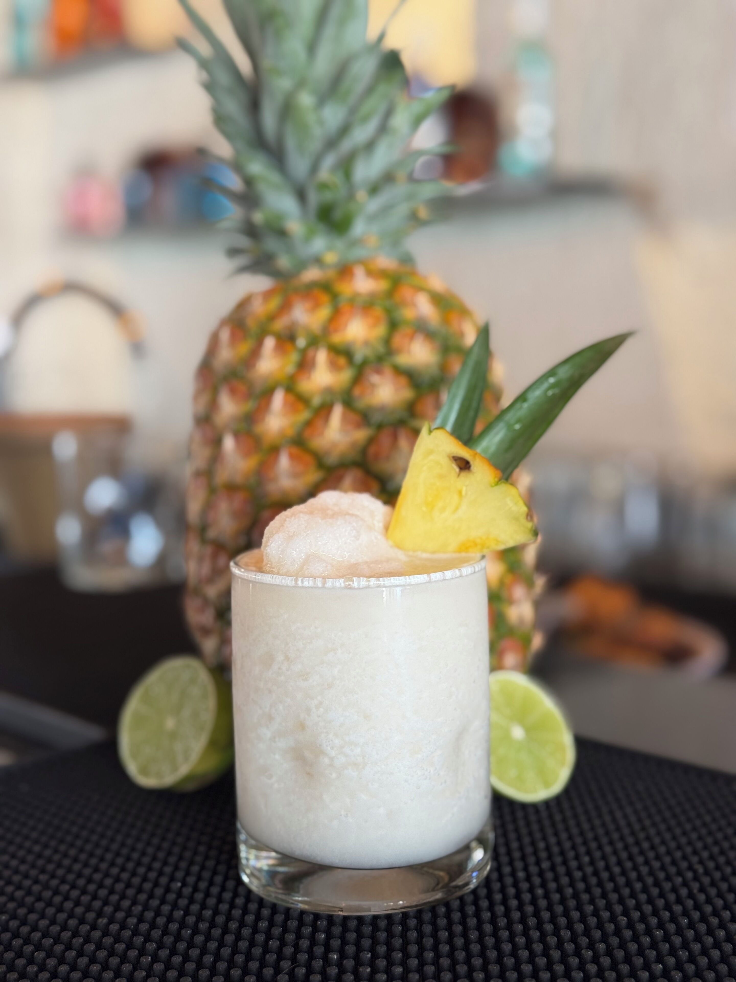 A glass of a frothy beverage with a pineapple slice garnish, lime wedges, and a pineapple in the background.