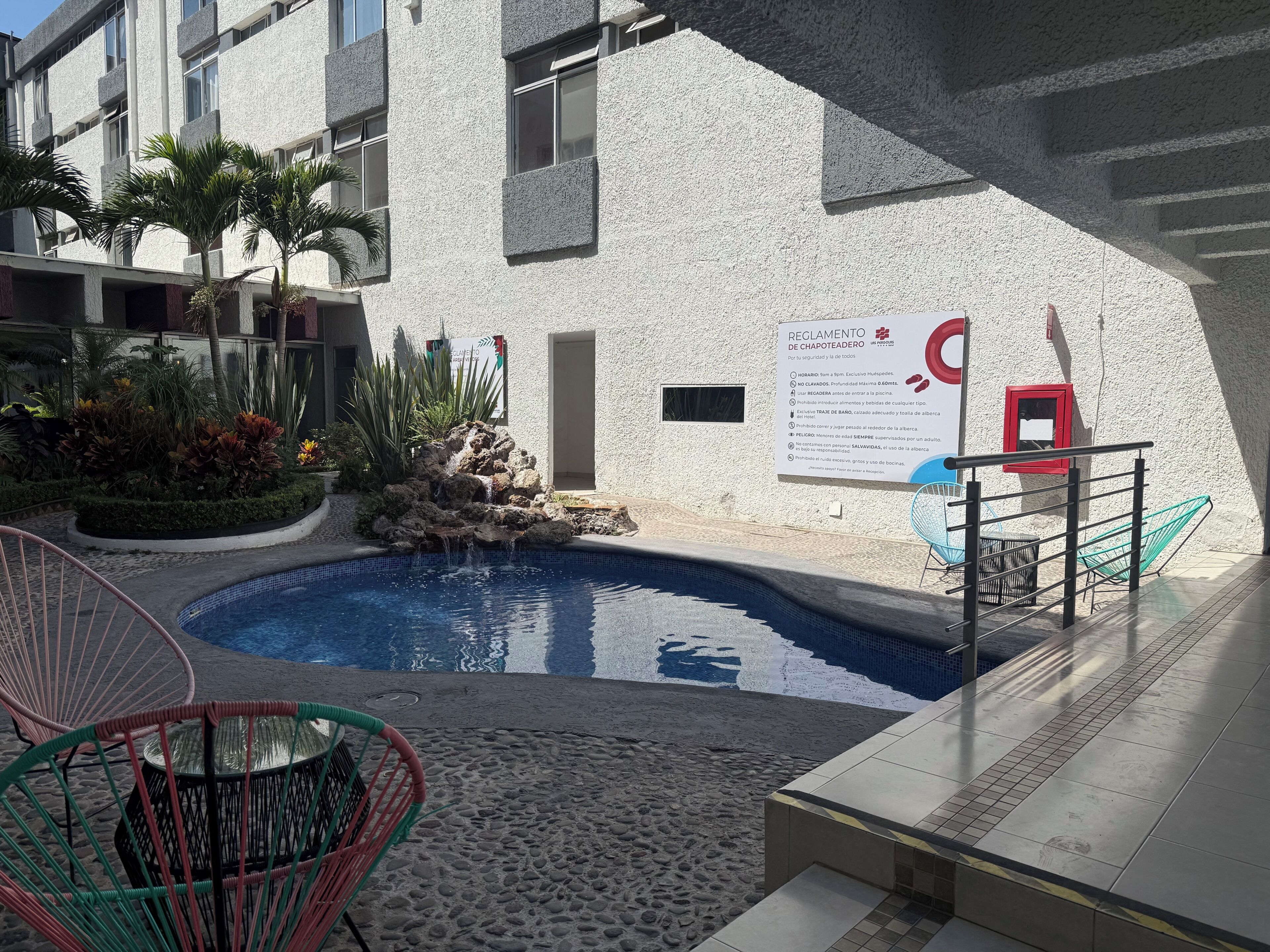 Una piscina con una formación rocosa, rodeada de sillones de descanso y un cartel con normas.