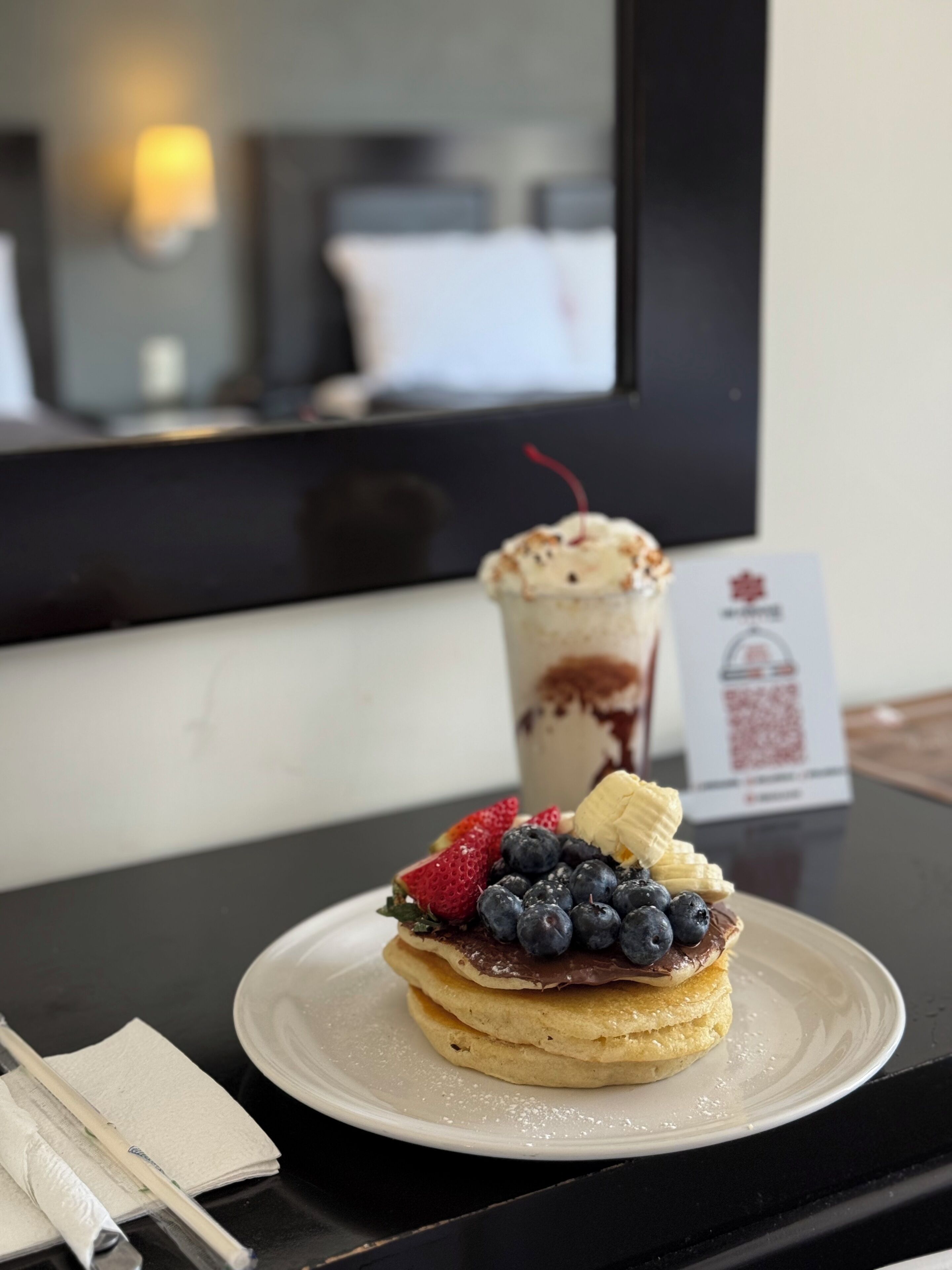 Een dessert op een bord met aardbeien, blauwe bessen en plakjes banaan, een drankje met slagroom, en een bord met een QR-code op een tafel.