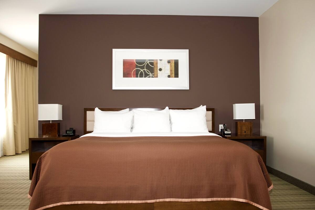 A hotel room with a large bed, two bedside tables with lamps, a framed abstract artwork above the bed, and a window with curtains.